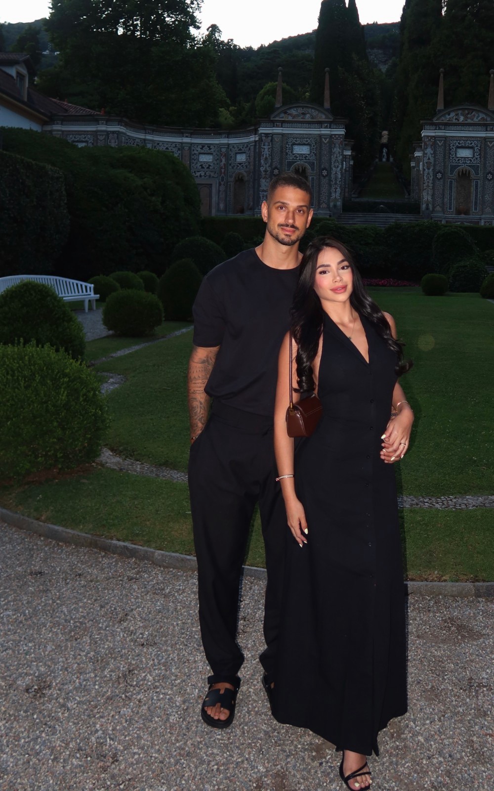 Valentina and Thyll, both dressed in black, standing together in a garden at dusk